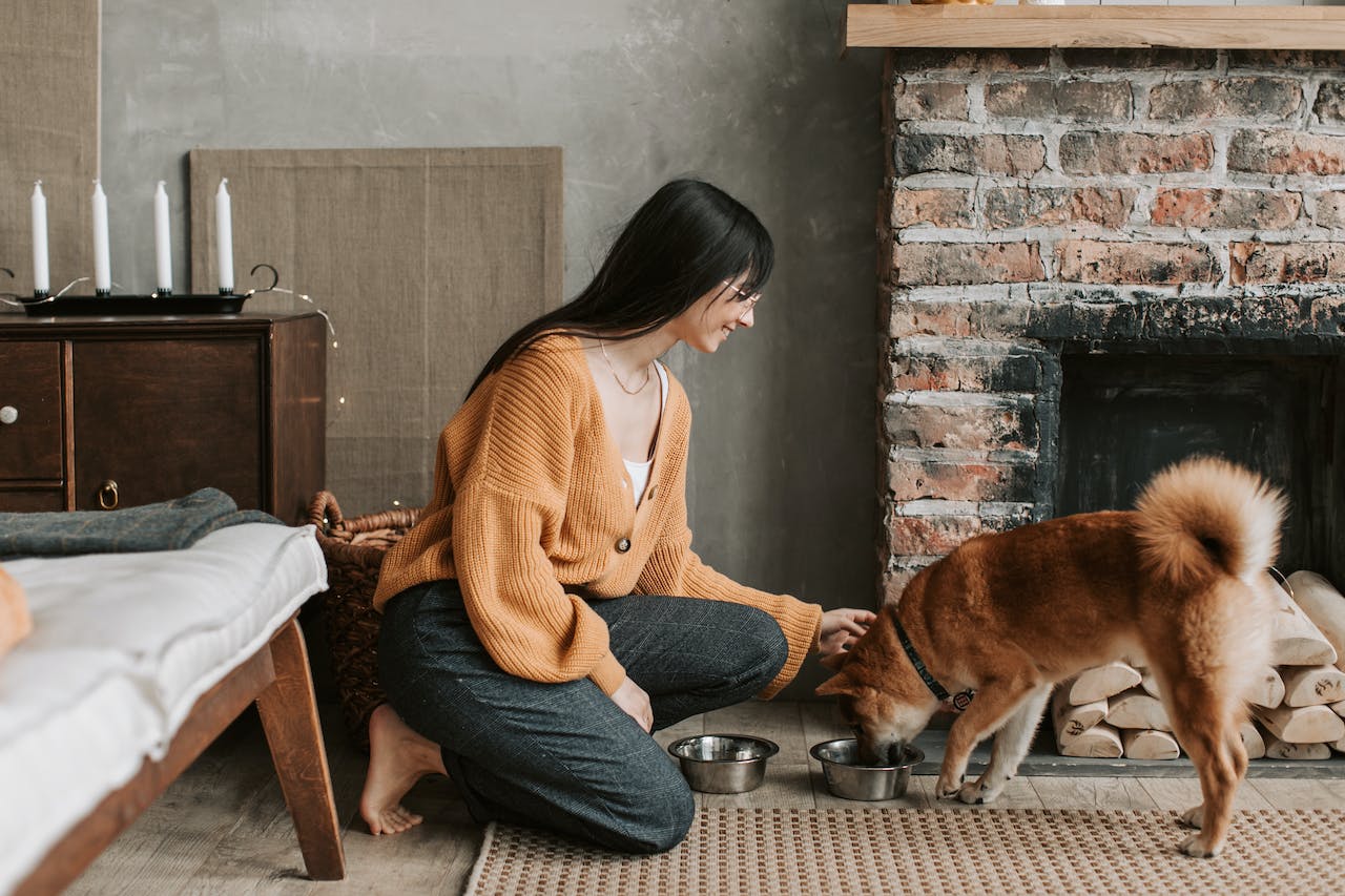 A woman in an orange sweater and jeans kneeling next t a tan dog eating from silver bowl
